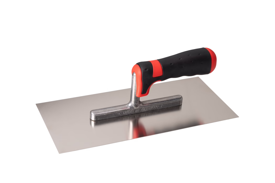 Pajarito_Dummy_Trowel soft handle Performance Line_034664_2021-02 A rectangular stainless-steel plastering trowel with a smooth flat blade and a short metal shank supporting an ergonomically shaped black and red soft-grip handle. The polished blade is reflective and shows a faint reflection of the handle; the metal shank is stamped MADE IN GERMANY. The handle has red accents at the front and rear and a textured rubber surface for a secure grip. The tool is photographed isolated on a white background from a slightly elevated side angle. Suitable for applying and smoothing mortar, plaster or render.
